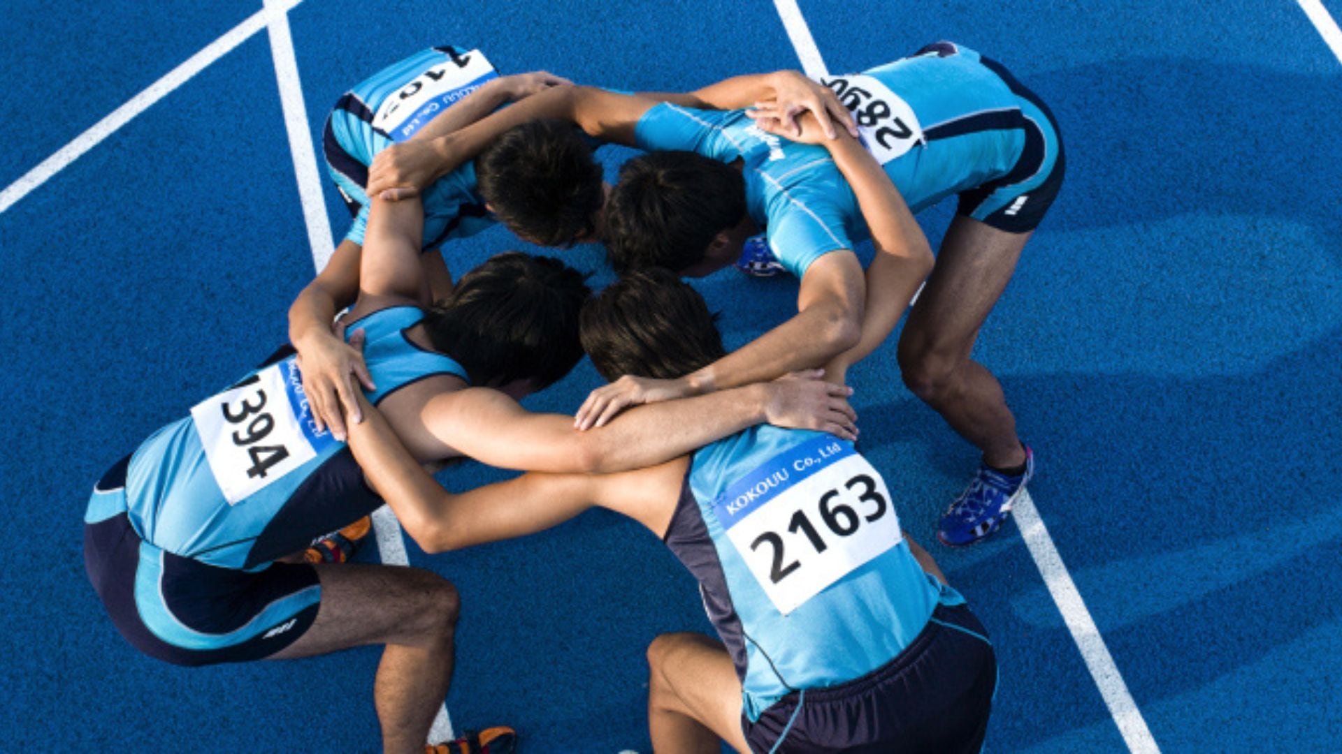 Four relay runners huddle on blue track preparing for race start