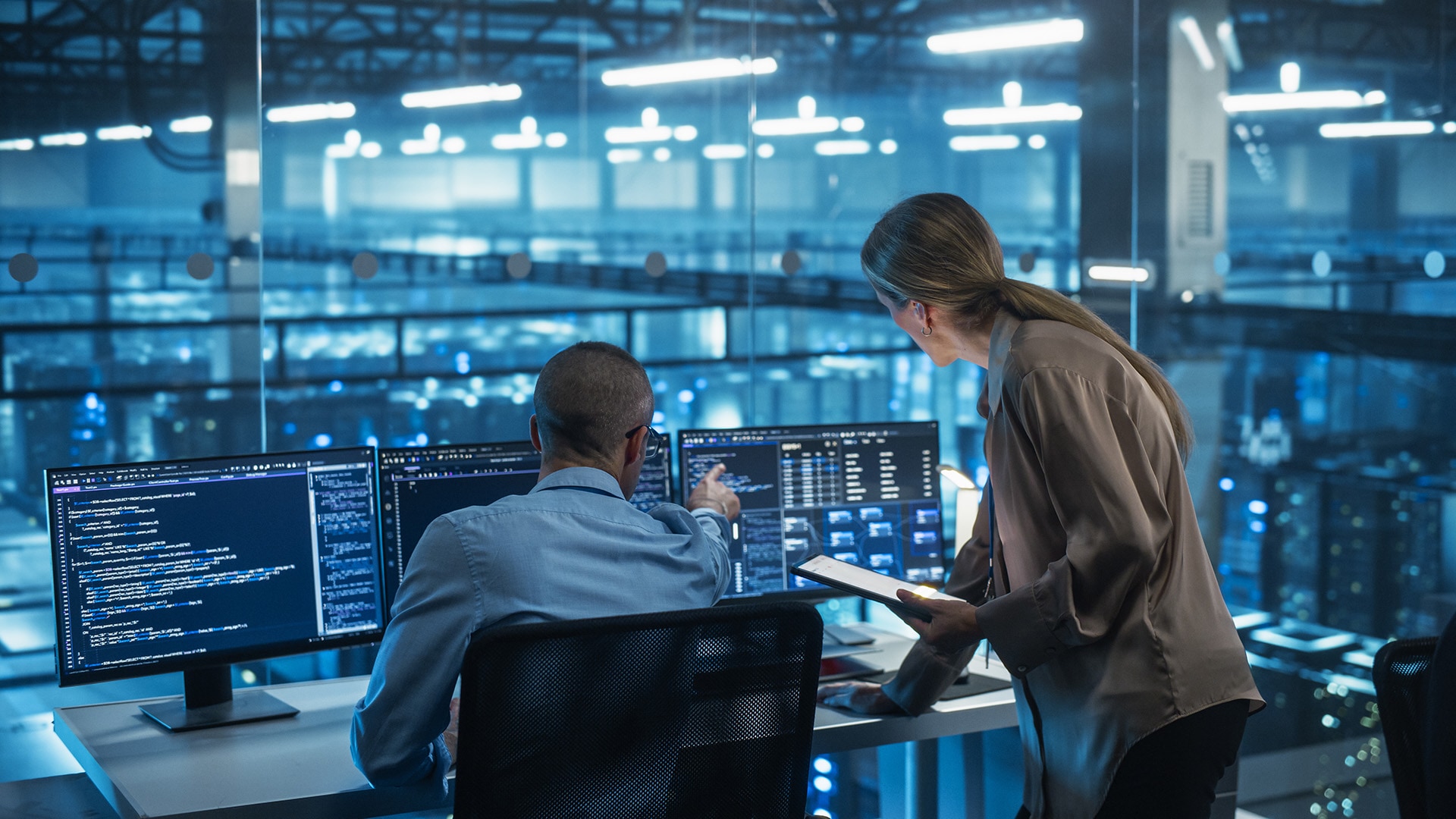 Two engineers reviewing code on multiple monitors in data center control room with servers in background