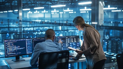 Two engineers reviewing code on multiple monitors in data center control room with servers in background