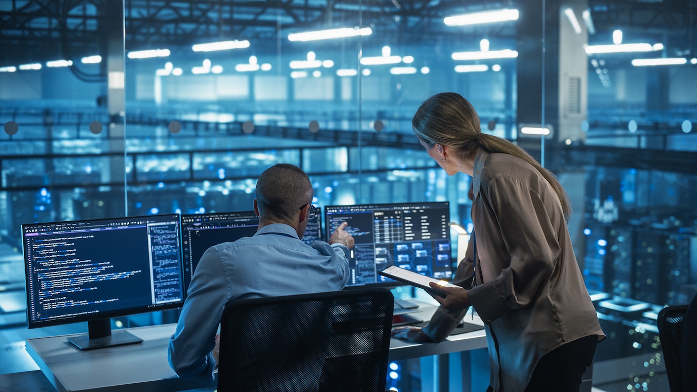 Two engineers reviewing code on multiple monitors in data center control room with servers in background