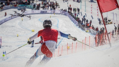 Skier racing downhill on snowy course with drone filming and spectators in background