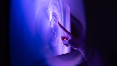 Finger touching glowing interactive digital surface in purple-blue light.
