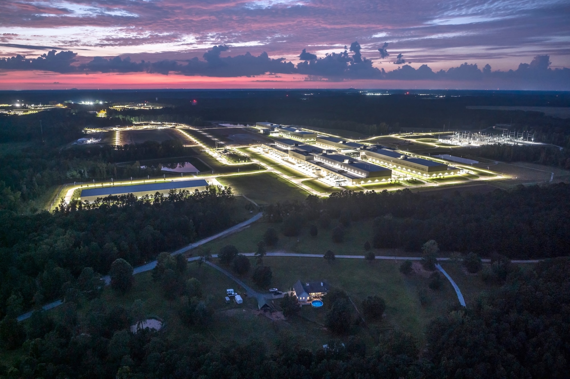 Aerial view of illuminated industrial campus at dusk surrounded by forest and rural landscape.