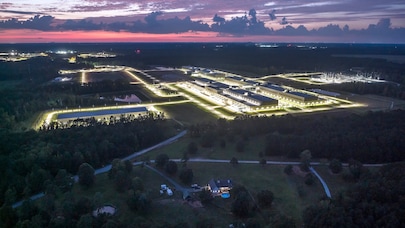 Aerial view of illuminated industrial campus at dusk surrounded by forest and rural landscape.
