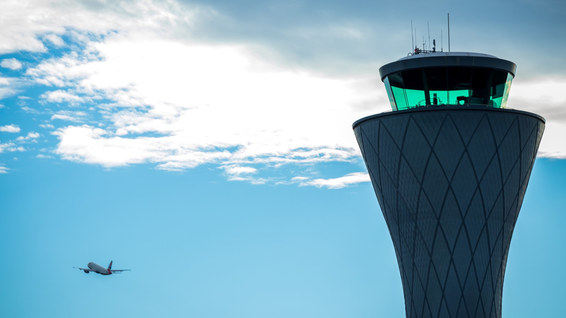 Air traffic control tower in the foreground with plane in the air.