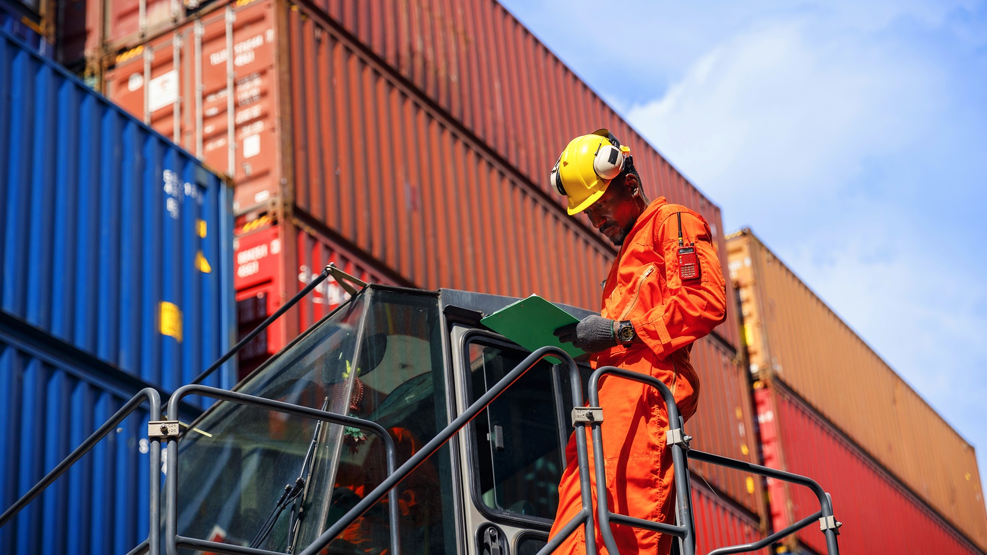 Man in hardhat checking info on clipboard in front of shipping containers