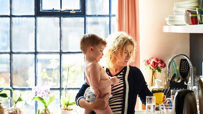 A mother holding a child in the kitchen of an ordinary home while IoT is working quietly in the background, unseen.