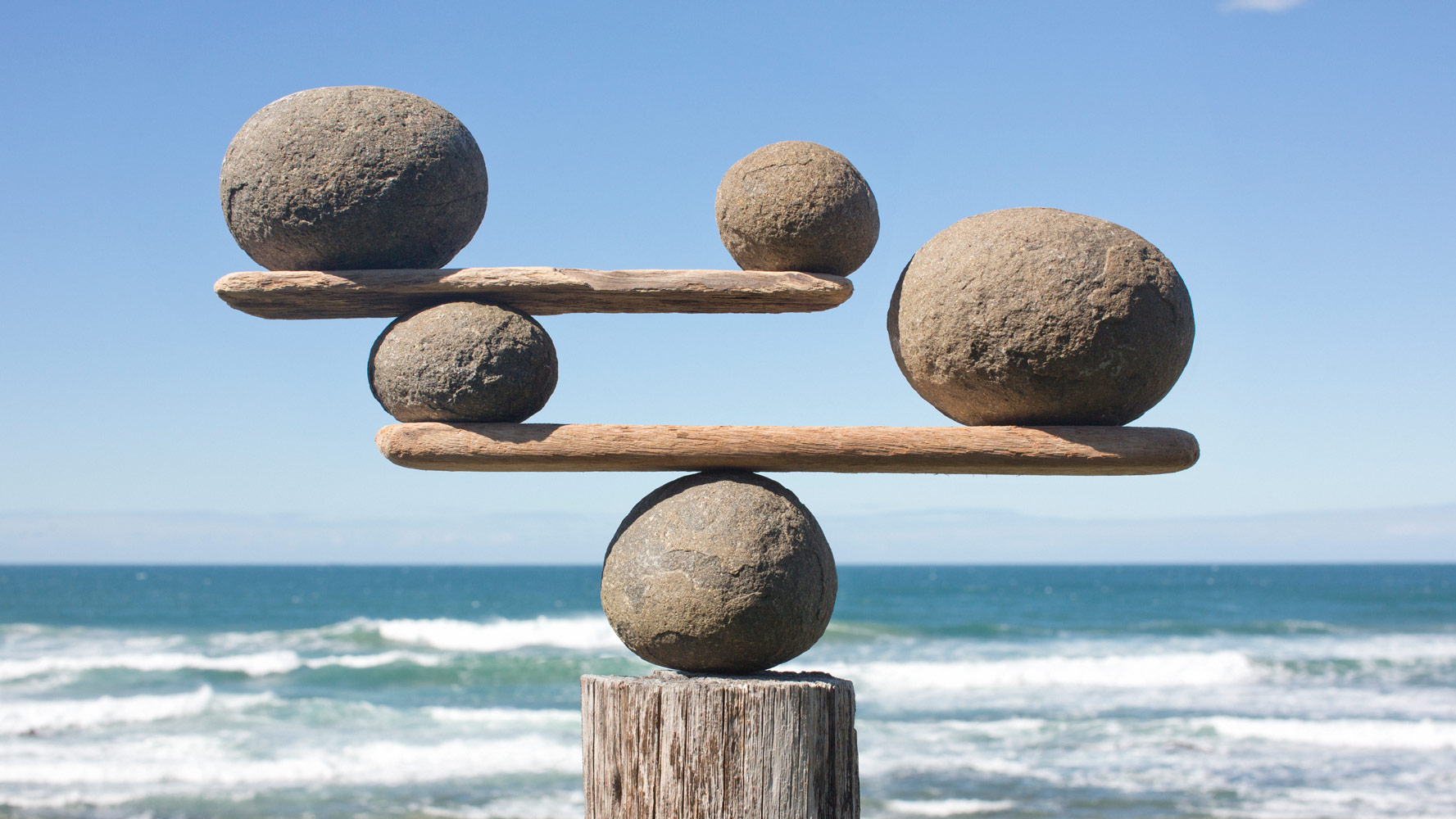 Balanced rocks and driftwood forming a precarious sculpture on a wooden post, with ocean waves in the background.