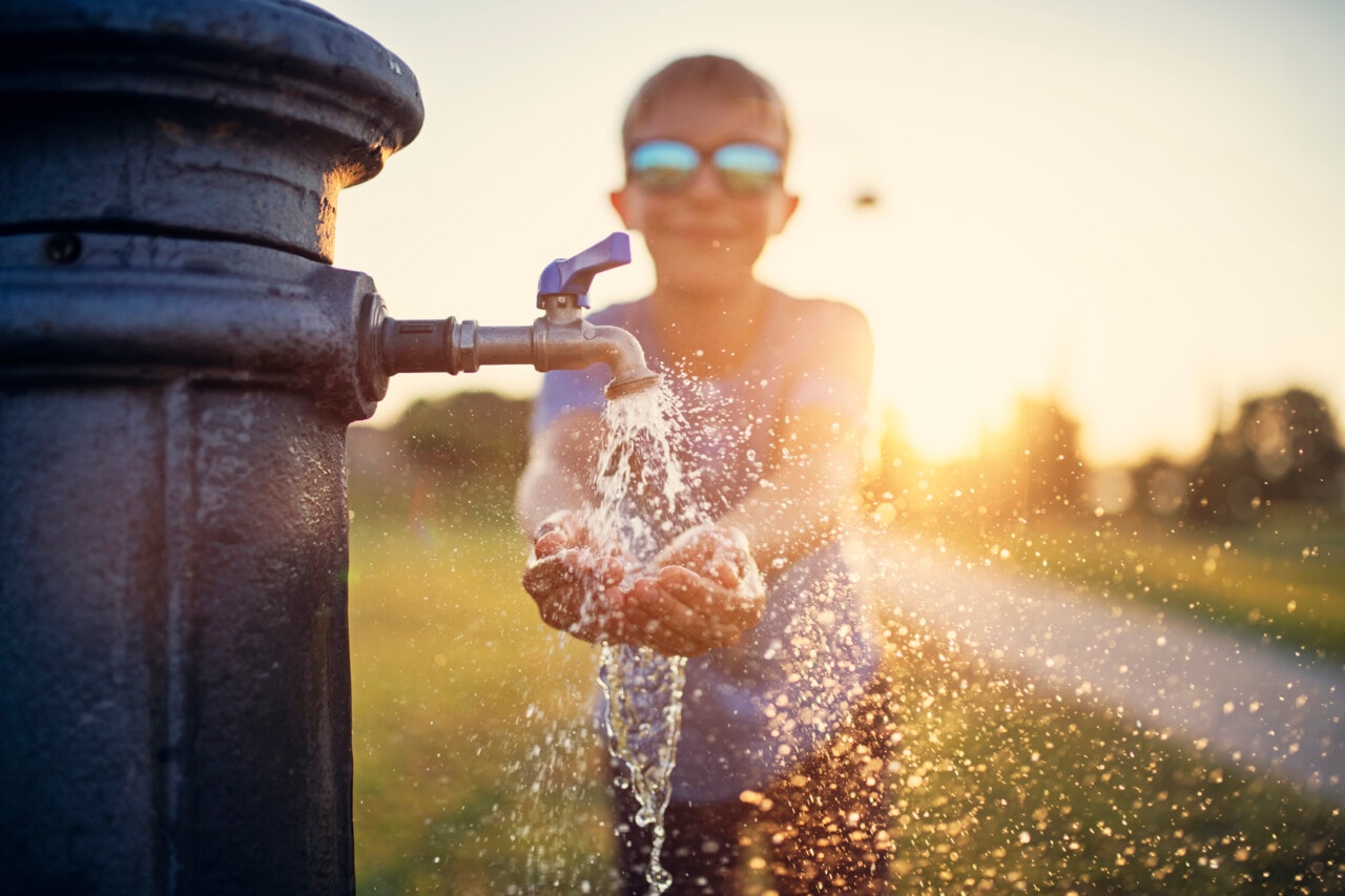 Child with sunglasses splashing water from outdoor tap at sunset, creating a sparkling spray effect.