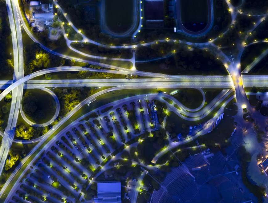 An aerial night view of a complex highway interchange or intersection. The roads are illuminated, creating striking patterns of light against the dark background.