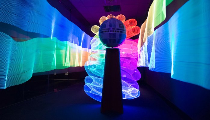 A model of the Telstar satellite, a dark spherical object with visible panels, sits atop a pedestal in a dimly lit room. Behind it, vibrant multicolored light installations create waves and patterns, including a fan-like array of illuminated circles.