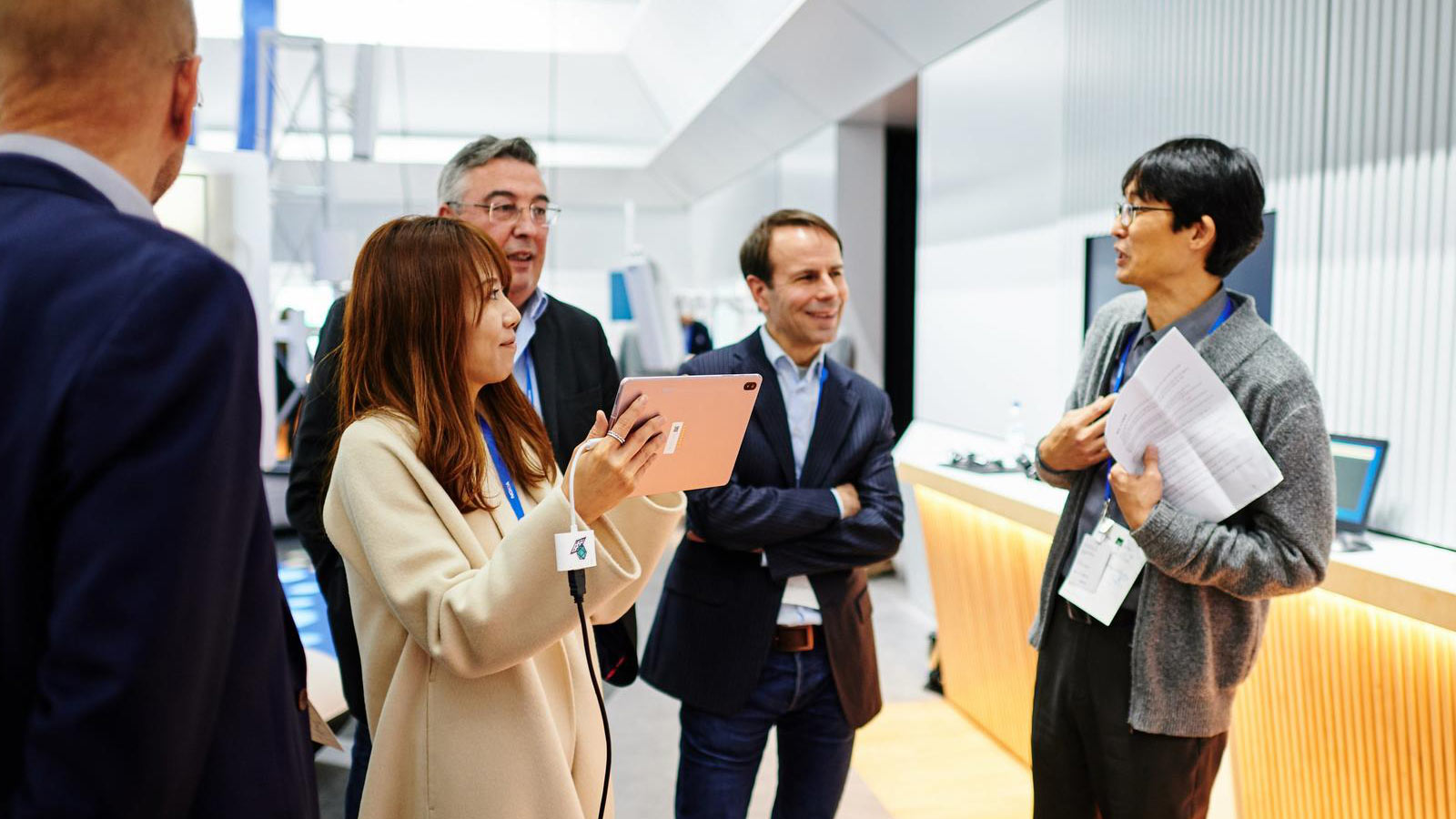 A group of four people, likely colleagues, engaged in a discussion. A woman is holding a tablet and appears to be showing something to the group.