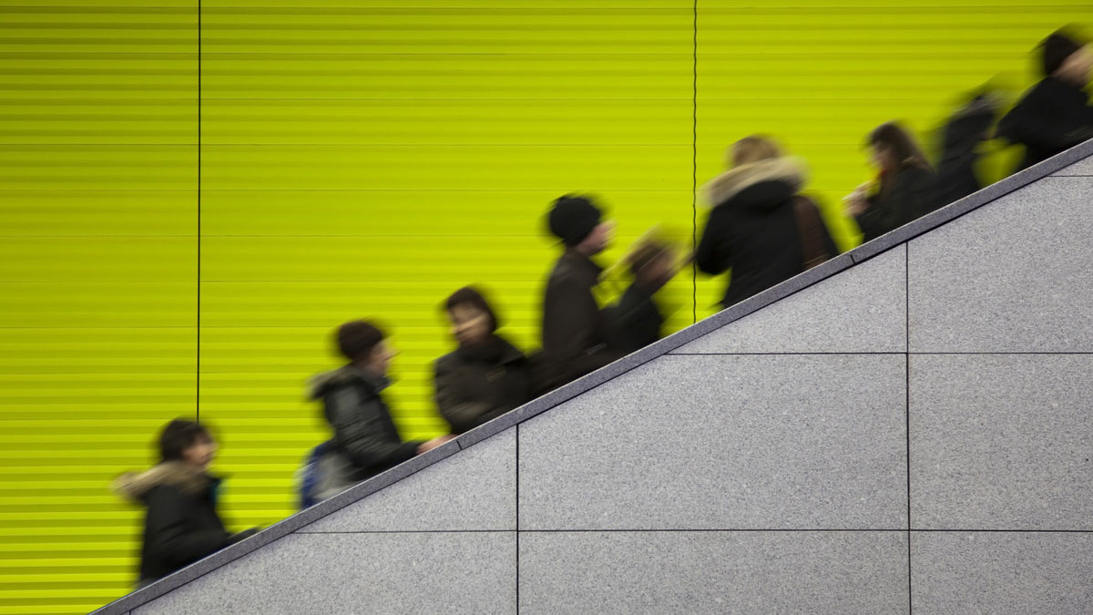 A group of people ascending a staircase or escalator, captured with motion blur. They are silhouetted against a bright lime-green background, likely a wall or panel.