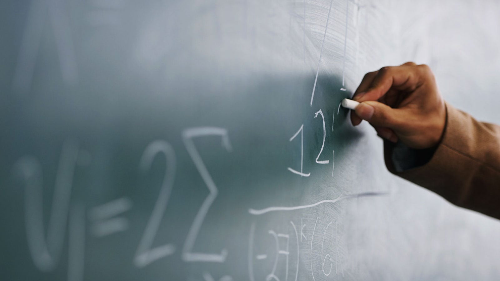 A close-up of a hand writing numbers in chalk on a dark green chalkboard.