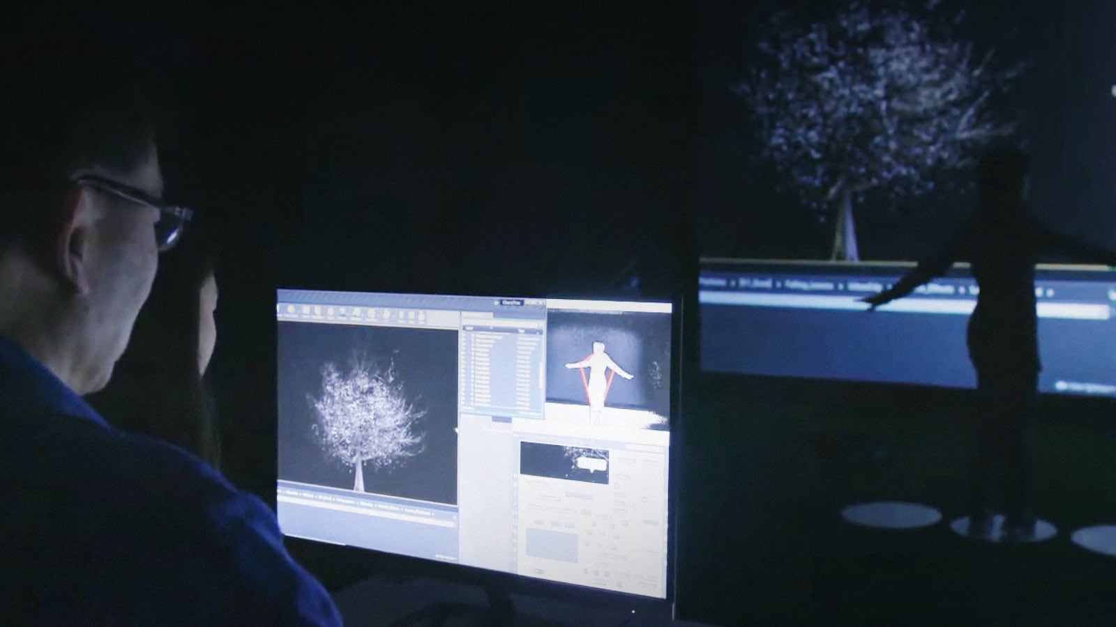 Person wearing glasses working on a computer, viewing 3D graphics software displaying a tree model and a human figure model on the screen.