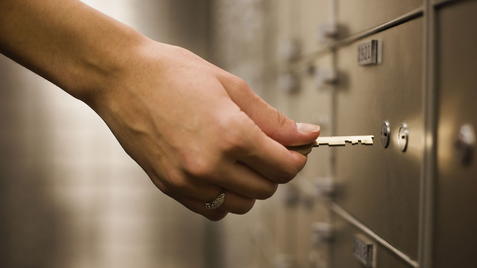 A close-up of a hand inserting a key into a safety deposit box. The hand is holding a small gold-colored key, poised to unlock one of many metal deposit boxes visible in the background.