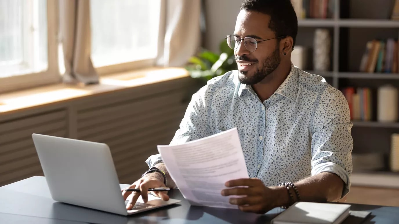 A smiling person holding papers in their hand and looking at their laptop screen.