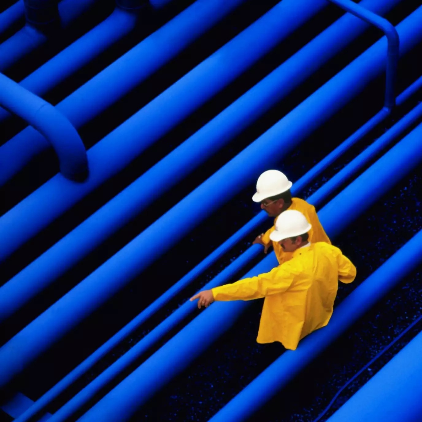 Two engineers in white hard hats and yellow jackets stand between blue pipes