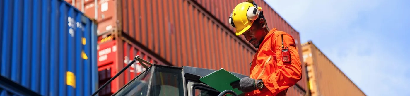Man in hardhat checking info on clipboard in front of shipping containers